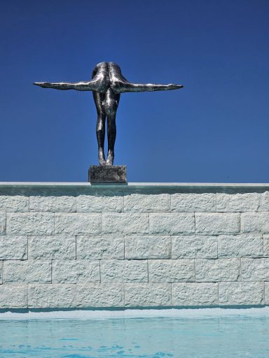 Skulptur eines ausgestreckten Menschen über einem Wasserspiegel, klarer blauer Himmel.
