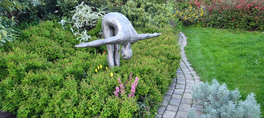 Skulptur eines sich biegenden Menschen in einem bepflanzten Garten.