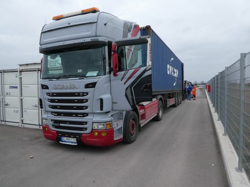 Ein Lkw mit einem blauen Container parkt auf einem schmalen Weg neben einem Zaun.