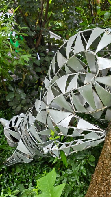 Skulptur eines katzenähnlichen Wesens aus einem glänzenden, geometrischen Material in grüner Umgebung.