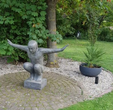 Sitzende, knieende Figur mit ausgebreiteten Armen in einem Garten.