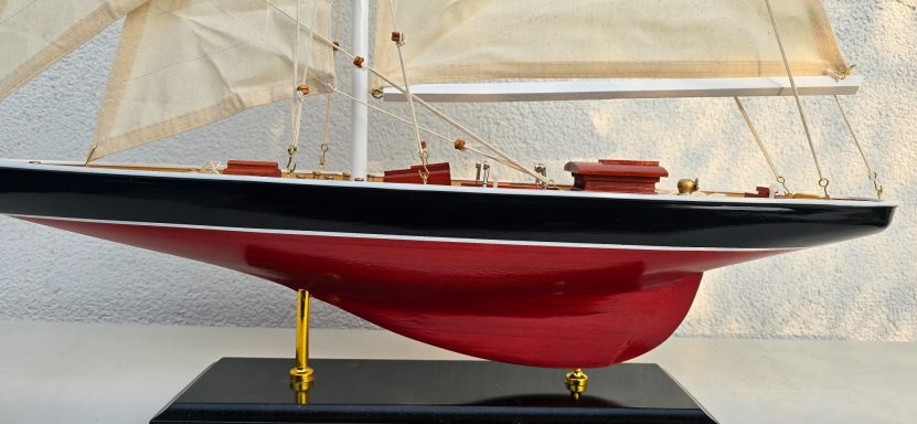 Modell eines Segelbootes mit rotem Rumpf und weißen Segeln auf einem schwarzen Sockel.