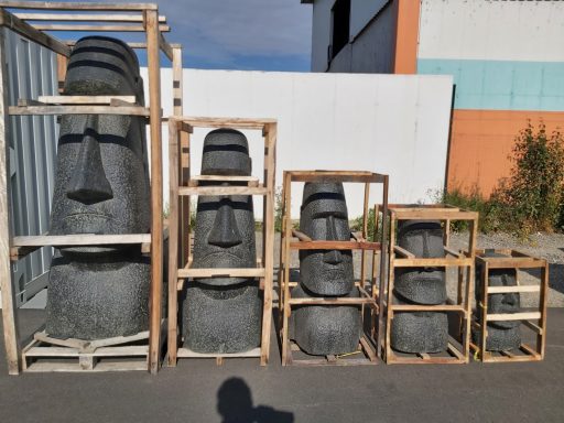 Sechs Moai-Statuen in Holzrahmen, unterschiedlich groß, auf einem Parkplatz.