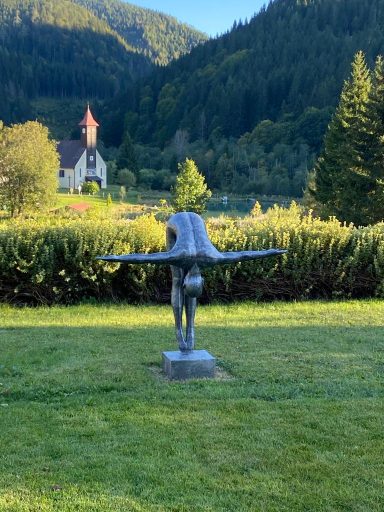 Bronze-Skulptur eines fliegenden Menschen vor einer Kirche in einer grünen Landschaft.