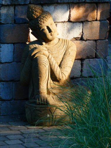 Steinerne Buddha-Statue in Ruheposition, umgeben von Ziegeln und Gräsern.