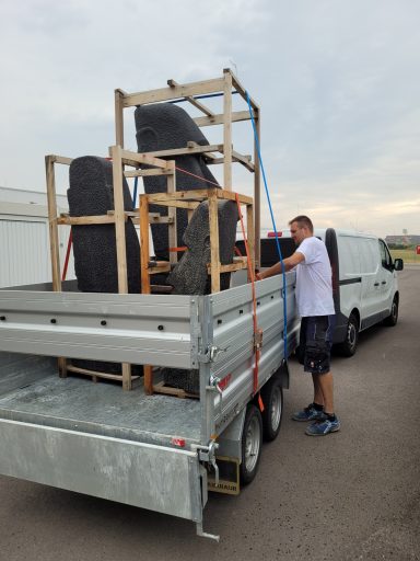 Ein Anhänger ist mit schweren Objekten und Möbeln beladen, ein Mann sichert die Ladung.
