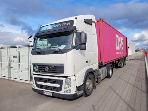 Weißer Lkw mit pinkem Auflieger auf einem Parkplatz. Wolken am Himmel.