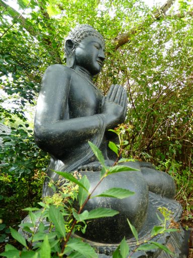 Ein 2m Hoher Buddha sitzt tief im Grün unter den Bäumen.