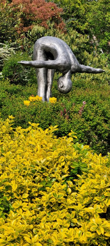 Skulptur eines sich bückenden Mannes, umgeben von bunten Pflanzen und Sträuchern.