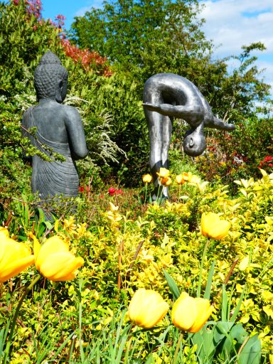 Skulpturen eines Buddha und einer gebogenen Figur umgeben von bunten Blumen.
