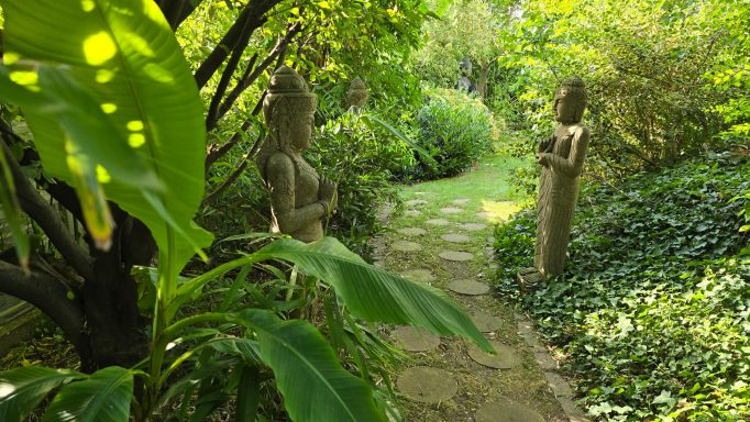 Ein steinern geformter Weg führt durch einen grünen Garten mit zwei Figuren.