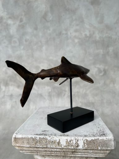 Bronze-Skulptur eines Hais auf einem schwarzen Sockel vor einem grauen Hintergrund.