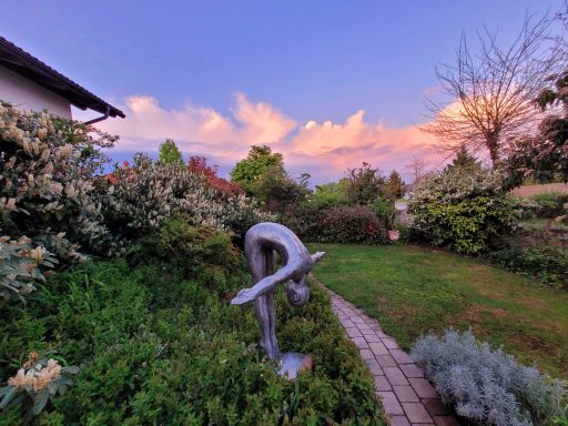 Bronze-Skulptur in einem Garten mit buntem Himmel im Hintergrund.