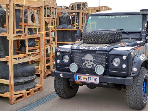 Schwarzer Geländewagen mit Totenkopfaufkleber und Holzpalette mit Skulpturen im Hintergrund.